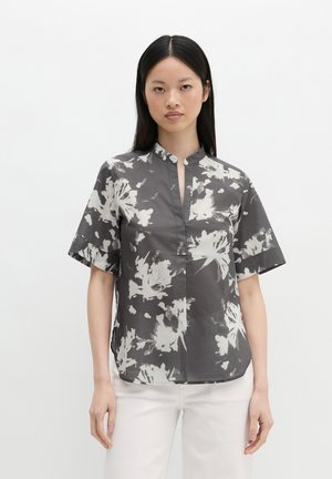 Vrouw met steil zwart haar draagt een grijze blouse met korte mouwen, wit bloemenpatroon en mandarijnkraag, gecombineerd met witte broek.