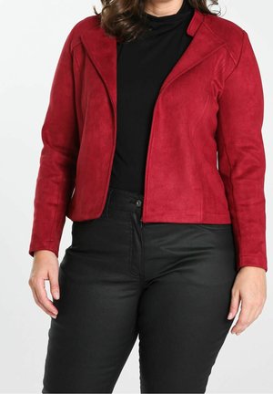 Donna che indossa una giacca aderente in camoscio rossa sopra una maglietta nera, abbinata a pantaloni neri in pelle a vita alta su sfondo uniforme.