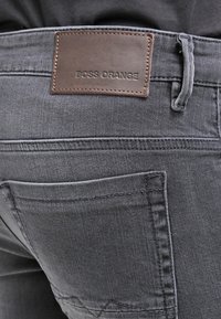 Mörkgrå jeans med en läderetikett som har texten "BOSS ORANGE" på midjebandet, med standard bakfickor och synliga sömnadsdetaljer.