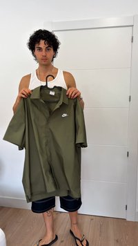 Olīvu zaļas krāsas īsu Piedurkņu pogājamā krekla, viegla materiāla, ar baltu Nike logo kreisajā krūtī, karājas uz melna pakaramā.