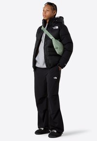 Joven con chaqueta y pantalones de plumas negros, sudadera gris, zapatillas negras sin cordones, bolso cruzado verde claro y gafas de sol en la cabeza.