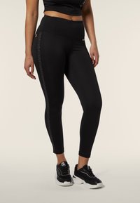 Leggings neri con vita alta, texture liscia, caratterizzati da un pannello laterale con un sottile motivo animale. Abbinati a scarpe da ginnastica nere.