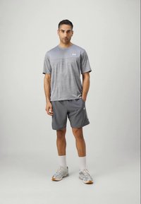 Mann steht, trägt ein graues Sport-T-Shirt und Shorts, weiße Socken und graue Laufschuhe, vor einem einfarbigen hellen Hintergrund.