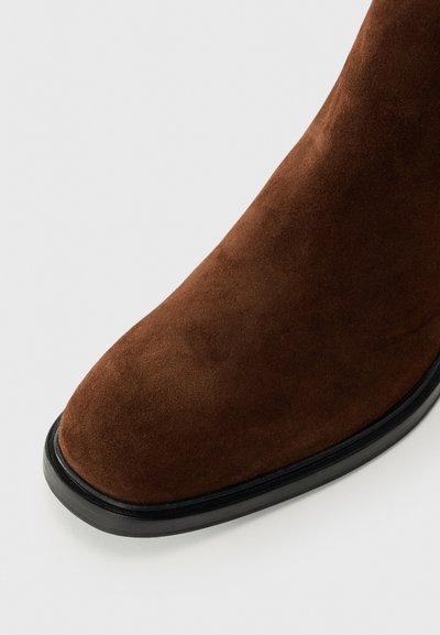 Botină de gleznă din suede maro cu vârf rotund și talpă din cauciuc negru, având o textură netedă și un design minimalist.