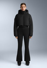 Czarna krótka kurtka puchowa z wysokim kołnierzem i zamkiem z przodu, w połączeniu z dopasowanymi czarnymi spodniami w dzwony i błyszczącymi czarnymi butami na platformie.