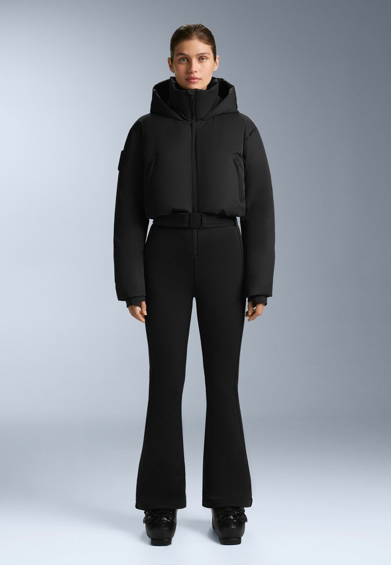 Czarna krótka kurtka puchowa z wysokim kołnierzem i zamkiem z przodu, w połączeniu z dopasowanymi czarnymi spodniami w dzwony i błyszczącymi czarnymi butami na platformie.