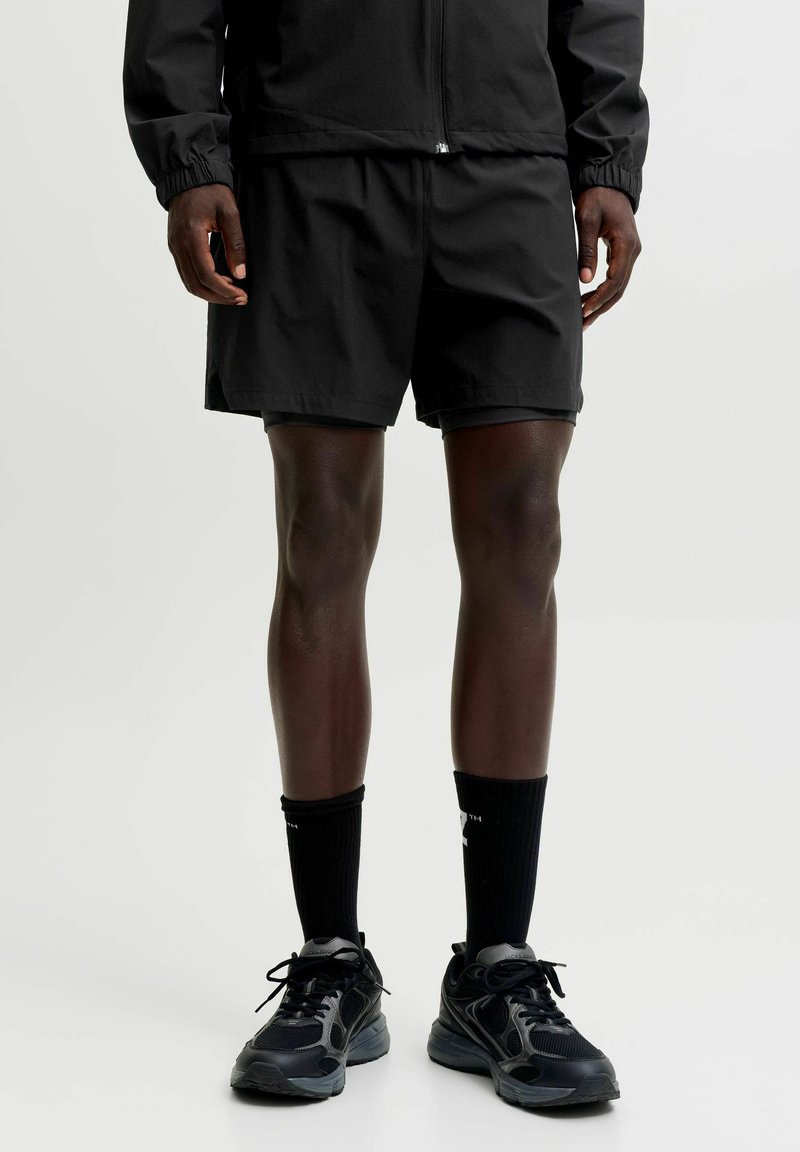 Personne portant un short de sport noir, une veste noire, des chaussettes noires mi-mollet et des chaussures de course noires, debout devant un fond neutre.