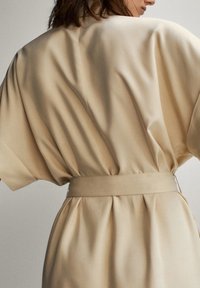 Beige mekko, jossa on lyhyet hihat, virtaava kangas ja vyö vyötäröllä. Selässä pehmeät laskokset ja hienovarainen laskostus, joka lisää tekstuuria.