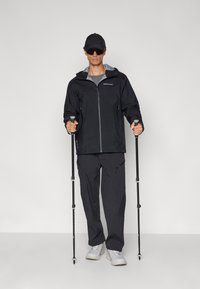 Veste noire imperméable avec des accents gris, poches zippées, associée à un pantalon noir et des bâtons de randonnée légers. Modèle portant une casquette et des lunettes de soleil.