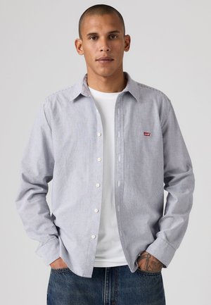 Homme aux cheveux courts portant une chemise légère à rayures boutonnée sur un t-shirt blanc et un jean bleu avec les mains dans les poches.