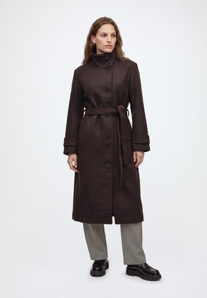 Femme portant un long manteau marron foncé avec des boutons et une ceinture à la taille, associé à un pantalon gris clair et des chaussures noires, se tenant devant un fond uni.