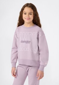 Niña joven con cabello largo y rizado, vistiendo un conjunto a juego de sudadera y pantalones morados claros de Champion, sonriendo con las manos relajadas a los lados.