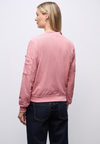 Donna con capelli biondi che indossa una giacca bomber in velluto a righe rosa e jeans blu scuro, in piedi con la schiena rivolta verso uno sfondo semplice.