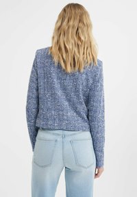 Blazer cropped blu textured con un motivo intrecciato, maniche lunghe e spalle strutturate, abbinato a jeans a vita alta blu chiaro.