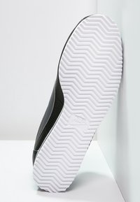 Baskets noires avec un matériau texturé et une semelle extérieure en caoutchouc blanc présentant un motif en zigzag. Le côté comporte un accent lisse noir et un détail de logo.