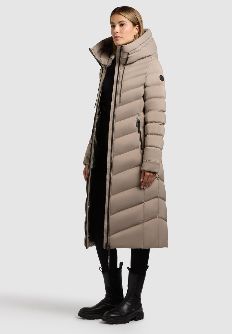 Lang beige dunjakke med chevron-quiltning, høj krave og lynlåslommer. Båret over sorte bukser og ankelstøvler, tekstureret stof.