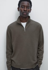Brun zip-up tröja gjord av mjukt stickat material med hög krage och subtil textur. Bärs över en vit T-shirt, uppställd mot en ljus bakgrund.