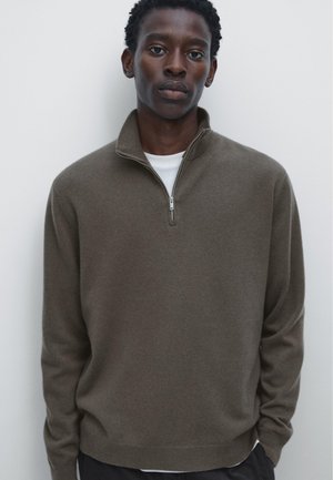 Brauner Reißverschluss-Pullover aus weichem Strickmaterial mit hohem Kragen und subtiler Textur. Getragen über einem weißen T-Shirt, präsentiert vor einem hellen Hintergrund.