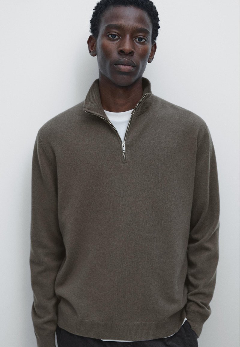Brun zip-up tröja gjord av mjukt stickat material med hög krage och subtil textur. Bärs över en vit T-shirt, uppställd mot en ljus bakgrund.