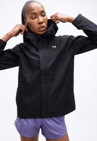 Femme ajustant le col d'une veste de pluie noire Under Armour, portant un short de sport violet Under Armour, sur un fond blanc.