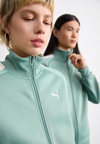Heledad rohelised lukuga spordijakid kõrge kaelusega, millel on valged aktsendid ja Puma logo. Sile tekstuur ja sportlik lõige.