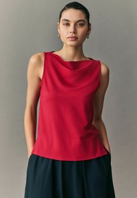 Czerwona bezrękawkowa bluzka z dekoltem w kształcie czołgu, gładką fakturą i lekko rozkloszowanym dołem, zestawiona z ciemną, zwiewną spódnicą.