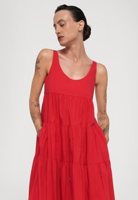 Abito rosso senza maniche realizzato in tessuto leggero, con un design a strati, scollatura ampia a girocollo e tasche laterali. Texture liscia in tutto l'abito.