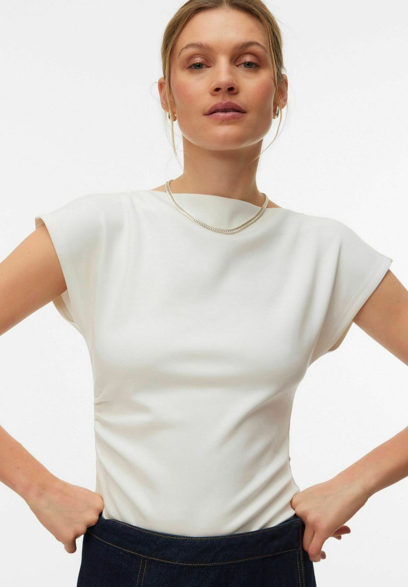 Haut blanc sans manches avec un col montant, coupe ajustée, texture lisse et fronces discrètes sur les côtés, assorti à une jupe en denim.