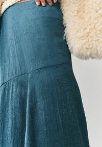 Gonna teal realizzata in tessuto testurizzato con una finitura increspata, dotata di vita aderente e fondo fluente. Uno strato esterno fluffy di colore crema è parzialmente visibile.