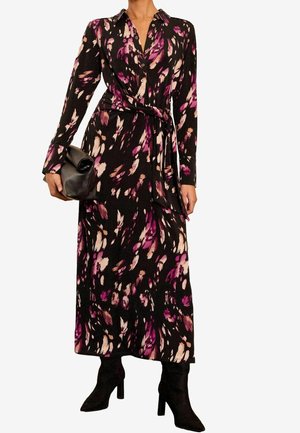 Femme portant une robe noire à manches longues et longueur cheville avec des motifs abstraits roses et beiges, tenant une pochette noire et chaussant des bottes à talons noires.