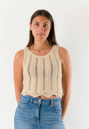 Donna con lunghi capelli scuri che indossa un top beige lavorato a maglia senza maniche e jeans blu a vita alta su uno sfondo bianco semplice.