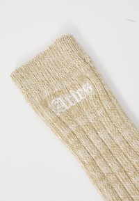 Strikkede sokker i lys beige med subtil tekstur. Har et hvidt broderet logo nær manchetterne. Cirkulær ribbenkant på toppen forbedrer pasformen.