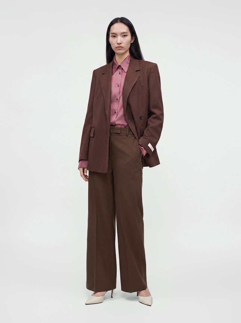 Woman wearing a dark brown suit with wide-leg trousers and blazer over a mauve button-up shirt and white pointed high heels, standing against white background.