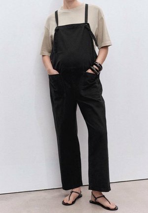 Osoba ubrana w luźne czarne ogrodniczki i beżową koszulkę z krótkim rękawem, z rękami w kieszeniach, nosząca czarne sandały na paski, stojąca na tle jednolitej ściany.