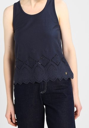Mouwloos marineblauw topje met geschulpte zoom en uitgestanst bloemmotief, gedragen met donkerblauwe spijkerbroek.