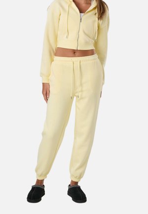 Kobieta w jasnożółtej krótkiej bluzie na zamek i pasujących spodniach dresowych, nosząca czarne buty wsuwane, na białym tle.