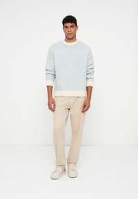 Fénykék kötött pulóver gyémántmintával, krémfehér bordázott nyakkivágással és mandzsettával. Bézs nadrággal és világos cipővel párosítva.