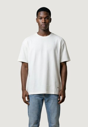 Giovane uomo con capelli corti che indossa una t-shirt bianca oversize e jeans azzurri, in piedi contro uno sfondo chiaro e uniforme.