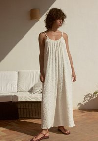 Robe blanche longue avec une apparence texturée et froissée, fines bretelles et coupe ample. Associée à des sandales plates rouges. Éclairage naturel.