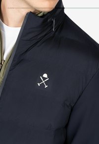 Hombre vistiendo una chaqueta acolchada azul marino con un logo de unos palos de polo cruzados en blanco y un casco en el pecho, superpuesta sobre un suéter verde con cremallera y una camisa blanca.