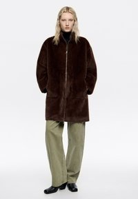 Abrigo de piel sintética marrón con un cuello alto, cierre de cremallera y bolsillos laterales, combinado con pantalones holgados verde oliva y zapatos negros.