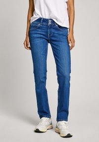 Vaqueros de mezclilla azul con un ajuste slim, cintura de media altura, dos bolsillos frontales y costuras en contraste. Llevados con una camiseta blanca y zapatillas.