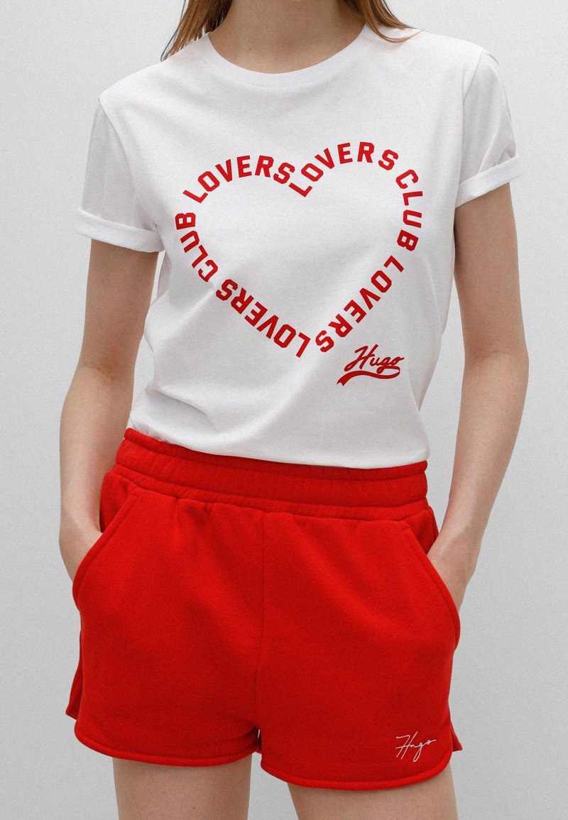 Hvid T-shirt med rød tekst, der danner et cirkulært mønster med teksten "LOVERS CLUB." Sammen med røde shorts med snoretræk og lommer.