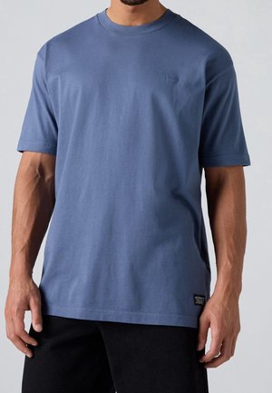Mand iført en ensfarvet blå T-shirt med korte ærmer og sorte bukser, stående foran en ensfarvet grå baggrund.