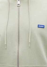Φούτερ με κουκούλα ανοιχτοπράσινο, ασημένιο φερμουάρ, κορδόνια που ταιριάζουν και μικρό μπλε ορθογώνιο έμβλημα με το λογότυπο "HUGO" στο στήθος.