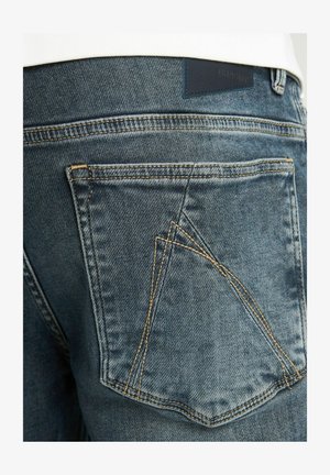 Denim jeans z večjo temno modro barvo, z vidnim šivom in značilno zasnovo žepov. Tekstuirana tkanina z rahlim bledenjem.