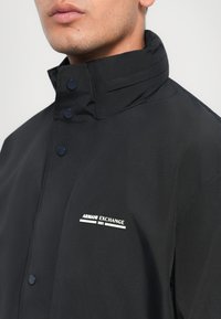 Black jacket featuring a high collar, button closure, and a white logo detail on the left chest. Made from smooth, lightweight fabric.