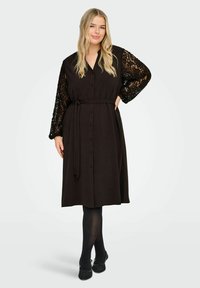 Czarna sukienka zapinana na guziki z paskiem w talii, ozdobiona koronkowymi detalami na długich rękawach i luźnym kroju. Noszona z czarnymi rajstopami i butami.