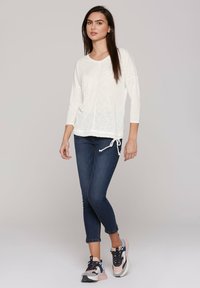 Weißes, leichtes Langarmshirt mit Kordelzugsaum, kombiniert mit dunkelblauen Skinny-Jeans und mehrfarbigen Sneakers.