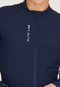 Giacca zip-up atletica blu con colletto alto, maniche lunghe e un discreto logo riflettente "ELITE LAB" sul davanti. Tessuto liscio e elasticizzato.